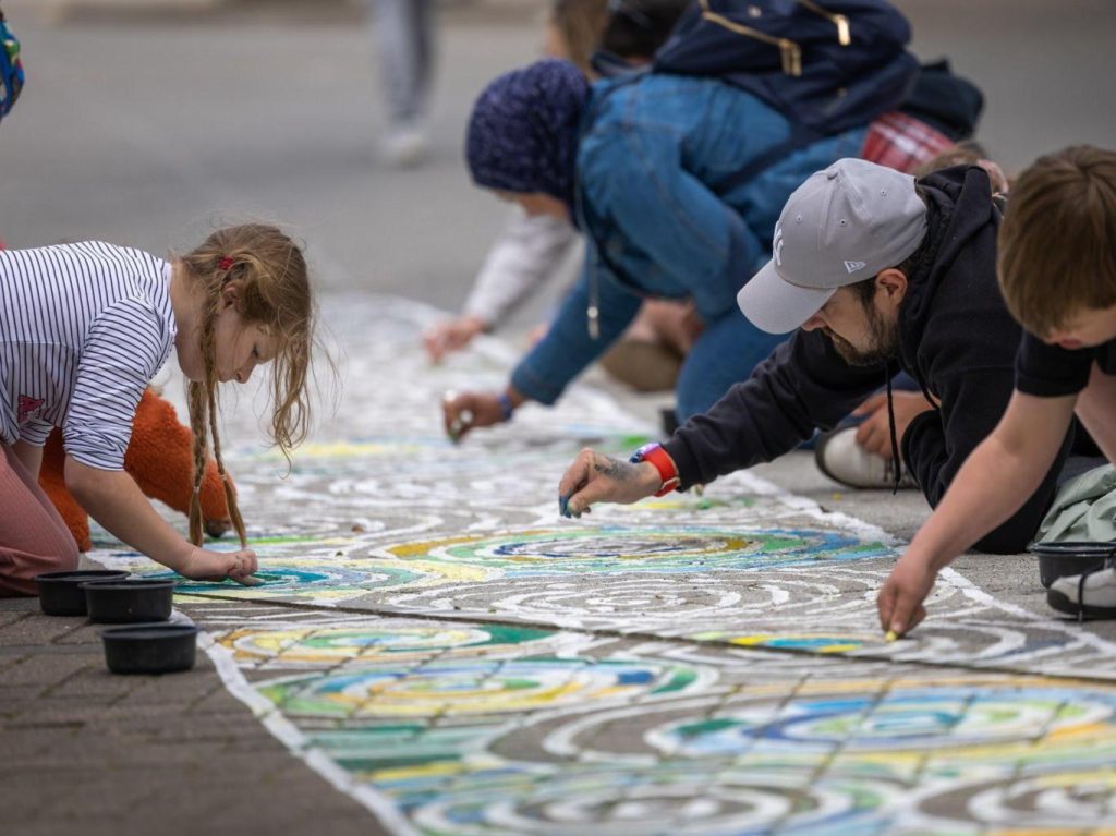People of all ages using chalk to create large-scale art on a pavement.